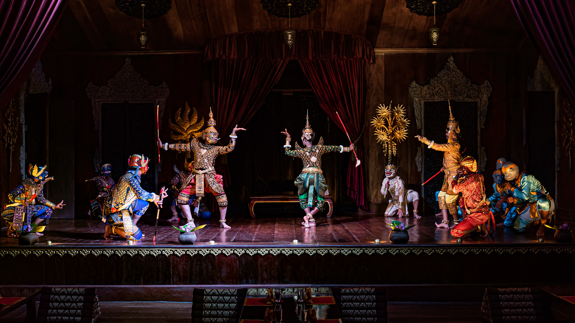 Performers wearing elaborate traditional costumes and masks dance on a wooden stage framed by dark curtains and ornate decorations at Apsara Theatre in Siem Reap, Cambodia