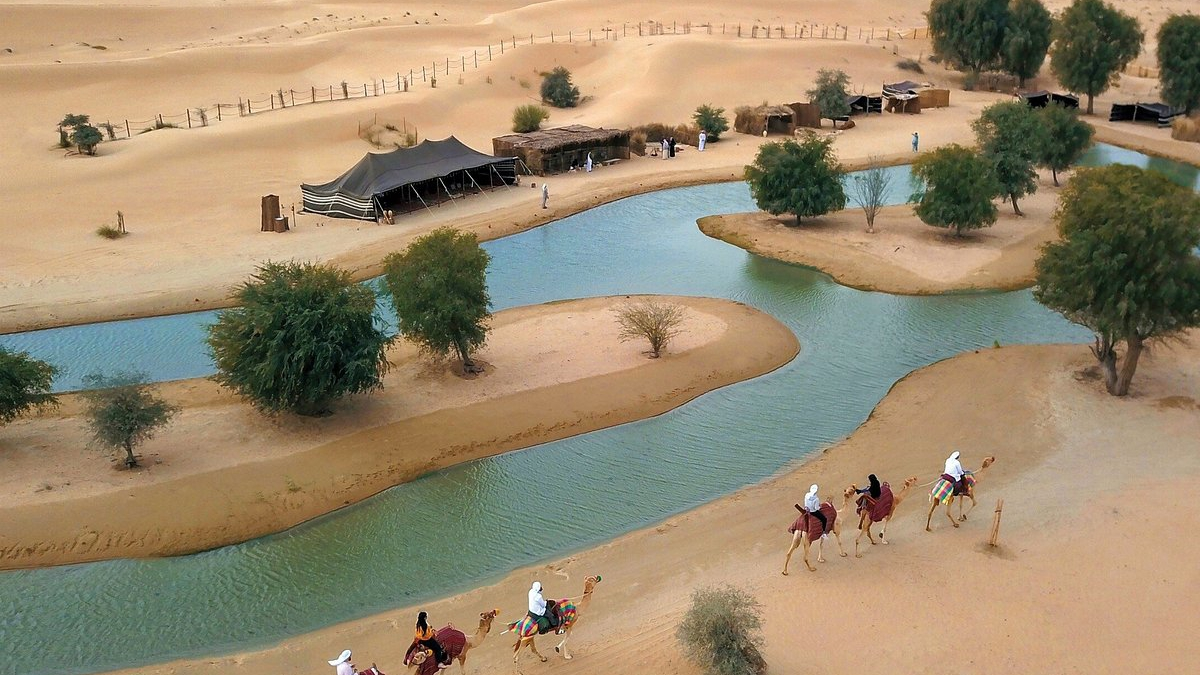 An oasis lake among the dunes of Al Marmoom from above, with a camel caravan on the sand